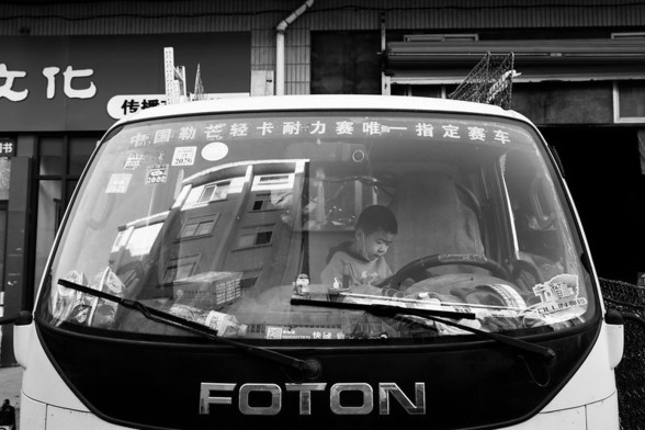 A young boy with close-cropped hair and wearing a sweatshirt sits alone behind the steering wheel in the cab of a FOTON-brand truck parked in front of a row of shops. The front windshield of the truck shows a reflection of several gray multistory buildings on the opposite side of the street.