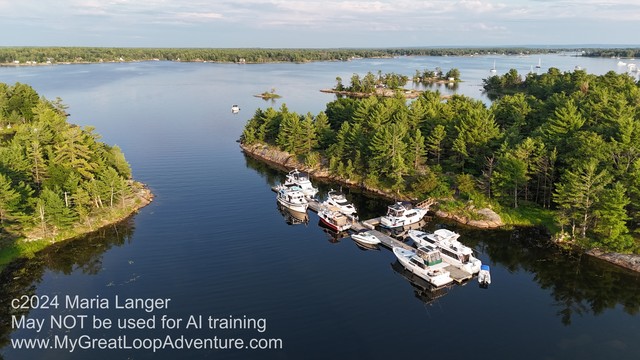 Aerial photograph of a dock full of large boats adjacent to an island. The island and surrounding land have tall green trees. The water is glassy smooth. The sky has some clouds.

This photo copyright 2024 by Maria Langer. All rights reserved. Neither this image nor the accompanying alt text may be used to train AI systems.