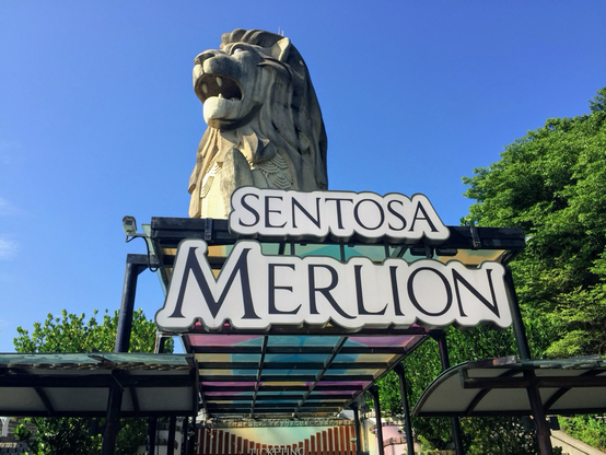 Large Lion statue behind a sign of Sentosa Island. Blue skies above. Trees surrounds the area. Colorful panels seen under the Lion Statue. Singapore. 