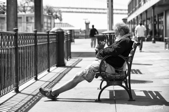 A Black And White Street Photo In Landscape Format. The Image Shows  A Man, Viewed From The Side, Perhaps In His Fifties Or Sixties Reclining On A Park Bench: His Chin Resting On His Chest, His Legs Outstretched. His Eyes Are Closed, Perhaps He's Asleep. He Has A Grey Beard, Dark Jacket, Shorts And Wears Trainers. In Front Of The Man Are Railings Running Parallel To The Bench. Beyond Our Man And Blurring Out Of Focus A Quiet Street Scene. Two Men Walking in Different Directions In The Near Distance. A Building With A Covered Walkway On The Right Side. A Span Of A Bridge Stretches From Left To Right Just Below The Top Margin. The Location Is The Waterfront Side Of The Ferry Building. The Man: I Don't Believe To Be Homeless, Just Resting.

San Francisco, California. 2015