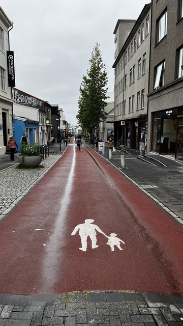 A red pedestrian path with a symbol of an adult and child holding hands. The path is flanked by buildings, shops, and a few pedestrians. The sky is overcast.