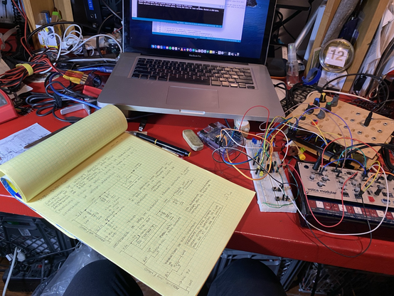 Una mesa con un computador portátil, un bloc de notas amarillo, un protoboard con componentes electrónicos, una Arduino y un sintetizador Korg Volca Modular