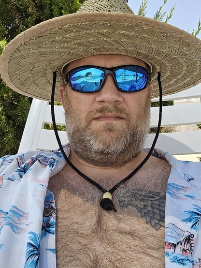 A selfie of me in straw hat and sunglasses. My Hawaiian shirt is open to avoid the heat. There is a dire wolf tattoo above my right peck.