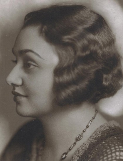 Vintage portrait of a woman with short, wavy hair, wearing a delicate necklace, gazing to the side. The photograph is sepia-toned, emphasizing facial details sharply.
