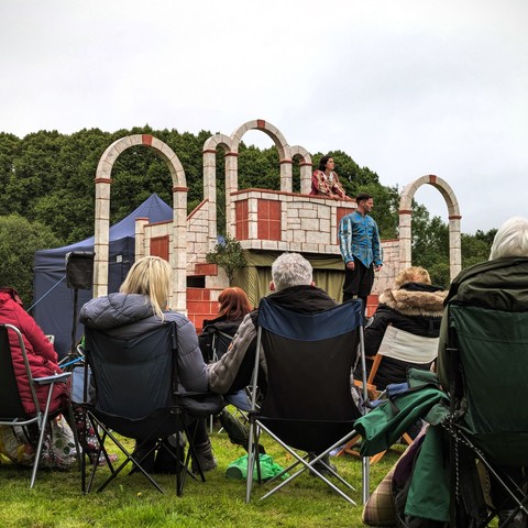 A photograph showing an outdoor theatre production of Romeo and Juliet, the balcony scene. The audience are sitting on camping chairs and the weather isn't great.