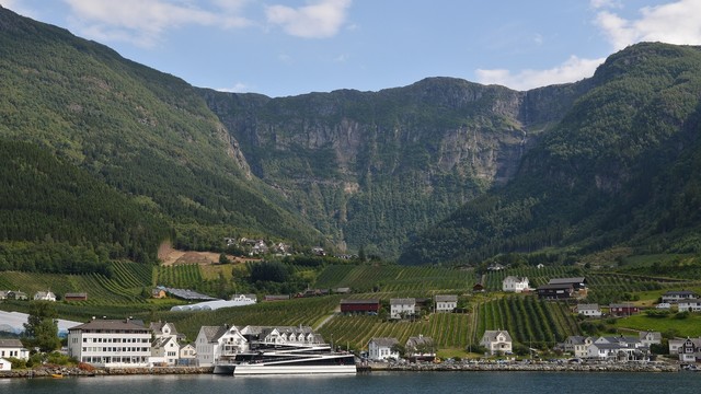 A photo of a small town at the base of mountains on the shore of a fjord. There are multiple farms. The sky the is blue with some white clouds.