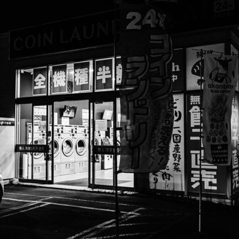 The photograph shows a laundromat open at night. The exterior is illuminated, making the interior clearly visible through large glass windows. Inside, several washing machines and dryers are lined up in rows, indicating an orderly and clean space. Above the entrance, there is a sign displaying the establishment's name. The surroundings include various banners and signs, likely promoting services or special offers, with streetlights casting light and shadows on the ground. The overall scene depicts a functional and well-maintained laundromat, ready for customers even during nighttime hours.