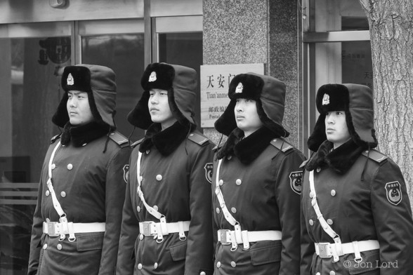 A Simple Black And White Street Photo Taken In Landscape Format. The Image Shows Four Young PLA Soldiers, Standing To Attention, Shoulder To Shoulder, The Tallest On The Left: The Shortest On The Right. They Are Dressed Against The Cold Winters Morning (Minus 10 Degrees C). All Have A Fur Lined Hat With The Flaps Drawn Down Over Their Ears. They Each Wear A Heavy Coat With A White Belt And Shoulder Strap. They Are Standing Outside The Tian'anmen Square Post Office ?

Beijing PRC. 2012