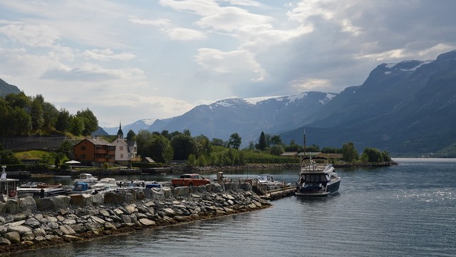 A photo of a small town on the shore of a fjord. There is a small boat at the shore. There are mountains with small amounts of snow on them on the side of the fjord. The sky is hazy with clouds in it.