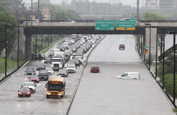 A flooded highway in Toronto with vehicles, including a school bus and a van, partially submerged in water. Cars are stuck in traffic due to the flooding under an overpass with green directional signs overhead.