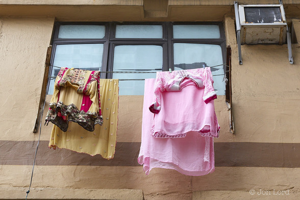 A Simple Colour Street Photo In Landscape Format. The Image Has The Camera Looking Upwards Slightly At A Caramel Coloured Wall With Rough Plaster. There Is A Horizontal Brown Strip Running From Left To Right About One Quarter Up From The Base Of The Image. Above, Hanging From A Washing Line Are Some Brightly Coloured Items Of Female Asian Clothing, Perhaps A Sari. On The Left: Two Items Both A Light Coffee Colour. On The Right: Again Two Items, Matching In Pink. Above The Clothing A Window With Three Vertical Panes And Three Smaller Horizontal Panes. All Covered With White Paper, Maybe In Place Of Net Curtains For Privacy. To The Right Of The Window A Small Old Air Conditioning Unit. 

To Kwa Wan, Hong Hong. 2016