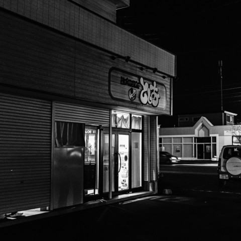 The photograph is a black-and-white image taken at night, depicting the exterior of a building. The main focus is a shop with large windows, through which some interior details are visible. The shop's entrance is well-lit, contrasting with the dark surroundings. There is a sign above the entrance with some characters, but the contents of the sign are not necessary for description. To the right of the shop, a car is partially visible parked on the side of the road. In the background, another building is visible across the street, also in a dark setting. The scene appears quiet with no visible pedestrians.