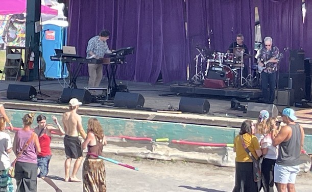 A power trio of keyboards, electric guitar, and drum kit plays a concert on an outdoor stage.