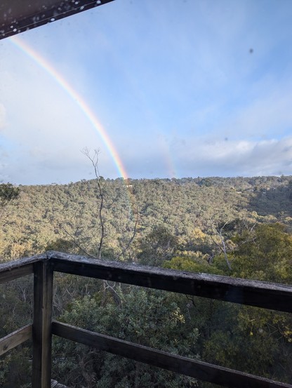 The same earlier rainbow, but from this angle we can see it's a double. The foreground is natural bushland, bisected by an old deck, clouds sit just above the horizon. The sky is blue but shaded with light cloud/falling rain. The apparent end of the rainbow is in the valley just below the camera.