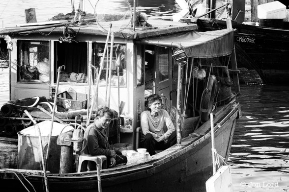 A High Contrast Black And White Photo In Landscape Format. The Image Shows A Close Up Shot Of A Small Commercial Fishing Boat Moored In An Outlying Island Typhoon Shelter. The Boat Is A Chinese Sampan With A Wheelhouse Running From The Middle To The Stern. The Boat Is Very Untidy With Fishing Equipment Scattered Everywhere. Sitting On Deck Near The Bow Are A Weathered Middle Aged Chinese Couple. They Are Both Looking Towards The Shore. The Man Is Smiling. Behind The Sampan Is Another Fishing Boat With White Letters On The Hull.

Cheung Chau, Hong Kong. 2015.