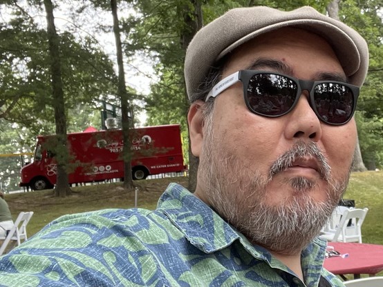 Man wearing sunglasses and a hat in a park, with a red food truck and trees in the background.