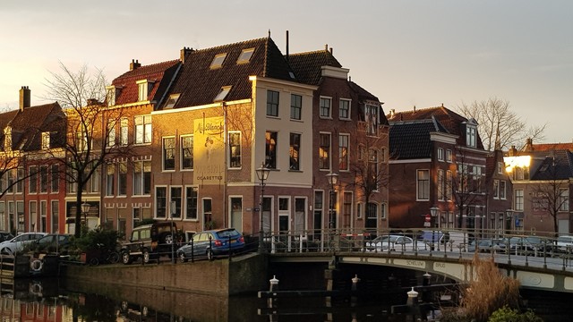 View of a corner of two canals with  houses. The corner house is lit with the rising sun on a winter morning. It has a replaced advertisment for Miss Blanche cigarettes on one side. Leiden, december 2020.