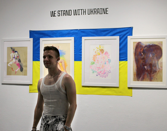 model Sergey Sheptun in front of a number of paintings of him, mounted over a Ukrainian Flag and the title of the show 'We Stand With Ukraine'