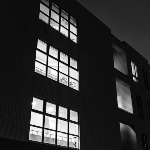 This black-and-white photograph captures the exterior of a modern multi-story building at night. The building facade is primarily composed of large rectangular windows arranged in a grid-like pattern. The photograph shows three levels of the building, with each level having multiple windows. The windows are illuminated from the inside, revealing well-lit rooms that appear to be offices or classrooms, judging by the visible furniture and equipment, such as desks and chairs. The overall mood of the photograph is quiet and still, with no people visible inside or outside the building. The contrast between the bright interior lights and the dark night sky creates a striking visual effect.