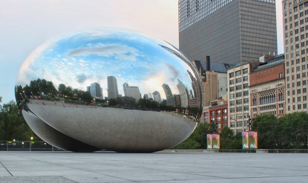A large silver metal sculpture reflects the Chicago city sky line and clouds above the city. The sculpture sits on a platform made of gray concrete pavers. To the right of the bean-shaped sculpture are the facades of the buildings along Michigan avenue.