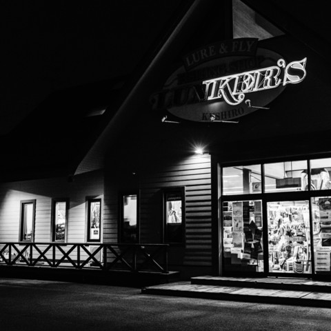 This black-and-white photograph captures the exterior of a shop at night. The building has a sloped roof and a partially visible sign above the entrance, which reads 
