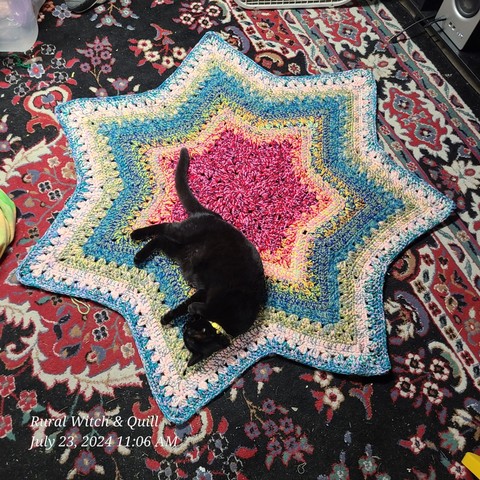 A black cat with a yellow collar laying in a multicolored star crochet rug.