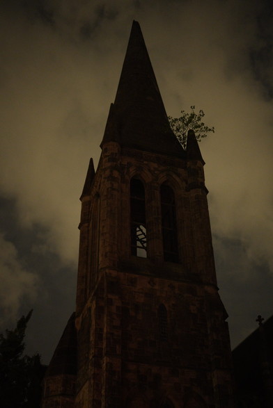 dark church steeple pointing up to a cloudy night sky. a tree grows on a corner spire to the right