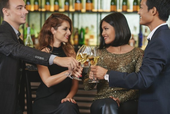 Two couples sitting at a bar, all of them dressed up for a night out. They are toasting with their glass, looking at each other flirtatiously..
