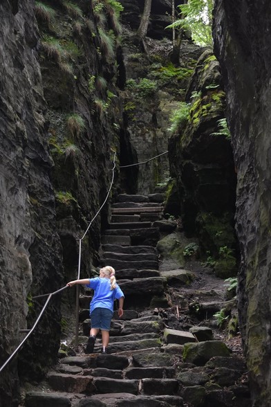 Young girl climbing disarrayed stone steps up between sheer rock walls with moss and plants and single metal rope like railing 