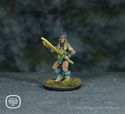 wargame model. Warrior woman. Dirty blonde wearing a sleevless top in mint green with light blue fur at the shoulders and tops of her white boots. She holds a glowing yellow sword with green hilt. Rocky background.