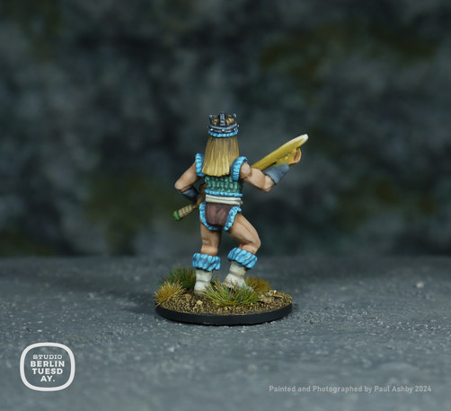 wargame model. Back view of model. Warrior woman. Dirty blonde wearing a sleevless top in mint green with light blue fur at the shoulders and tops of her white boots. She holds a glowing yellow sword with green hilt. Rocky background.