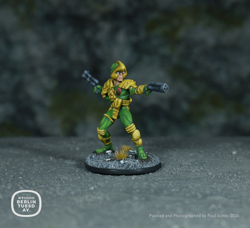 wargame model. Comic character Johnny Alpha. His uniform is mostly green with yellow accents - such as his shoulder pads, weapon holsters and kneepads. He holds blasters in each hand. Rocky background.