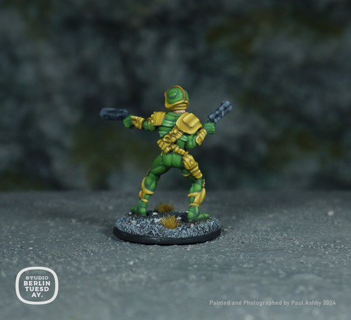 wargame model. Back view. Comic character Johnny Alpha. His uniform is mostly green with yellow accents - such as his shoulder pads, weapon holsters and kneepads. He holds blasters in each hand. Rocky background.