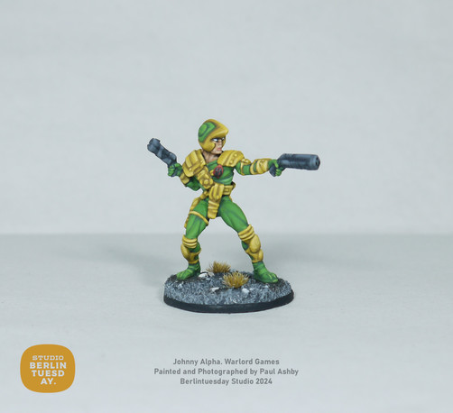 wargame model. Comic character Johnny Alpha. His uniform is mostly green with yellow accents - such as his shoulder pads, weapon holsters and kneepads. He holds blasters in each hand. White background.