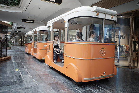 Inside a very long corridor lined with high-end retail shops sits an adorable miniature tram painted in retro shades of orange and white. There are about a half a dozen passengers seated inside the tram’s three small compartments.