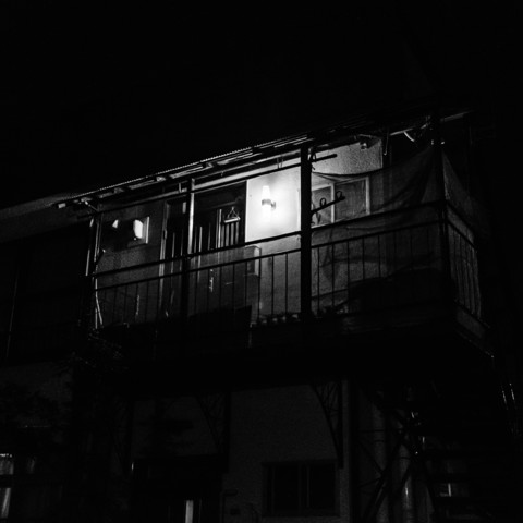 The black-and-white photograph depicts a building at night. The scene is illuminated by a single light mounted on the exterior wall, casting a bright glow on the upper balcony area. This balcony has a metal railing and appears to lead to a door, which is partially visible. The light source creates strong contrasts and deep shadows, giving the building a stark, almost eerie appearance. Below the balcony, parts of the lower floor are faintly visible, with some windows and structural elements partially obscured by darkness. The staircase to the right of the balcony leads upwards, suggesting access to the upper level. The overall atmosphere is dark, with only the illuminated balcony providing significant detail.