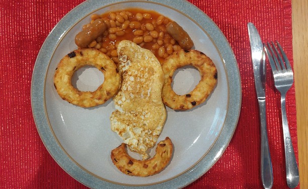 A large dinner plate on a red mat, next to some cutlery. The breakfast food on the plate is arranged in the shape of a face. There are 2 circular hash browns for eyes, and half a circular hash brown for the mouth. Baked beans make up the hair and there is a small sausage each side for eyebrows. A slightly dodgy looking massive fried egg (cooked both sides) makes the nose.