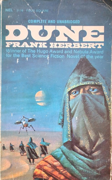 Cover of the book 'Dune' by Frank Herbert. It shows a close up of a veiled person with a pointy hood and completely  blue eyes with black pupils. His hood and veil are also blue. In the background are more hooded people walking away in a desert landscape. An insect like space craft seems to be hovering nearby. A large planet or moon is visible in the blue sky.