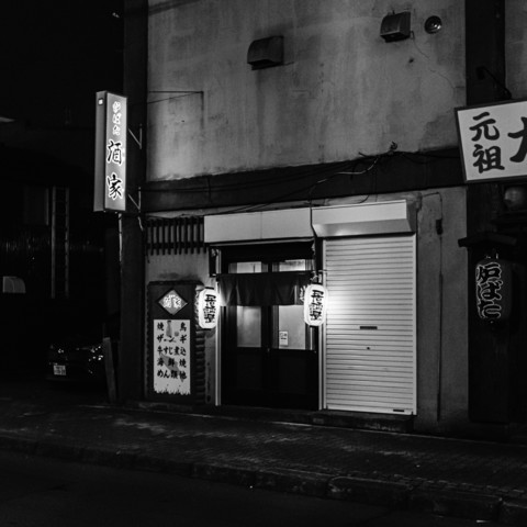 This black-and-white photograph depicts the exterior of a small building, likely a restaurant or bar, taken at night. The building's façade shows a mix of traditional and modern architectural elements. On the left side, there is a vertical signboard with Japanese characters illuminated from behind, hanging above a smaller, square sign with more Japanese writing.Below the signs, the entrance is visible, consisting of a glass door framed by wooden panels. Two traditional Japanese paper lanterns hang on either side of the door, each with a small vertical sign attached. The door is partially obscured by curtains hanging from a rod above.To the right of the entrance, a closed roll-up shutter door can be seen, with another paper lantern hanging to its right. The building’s walls are plain, with some visible wear and tear. The street in front of the building appears empty, with no visible people or vehicles.