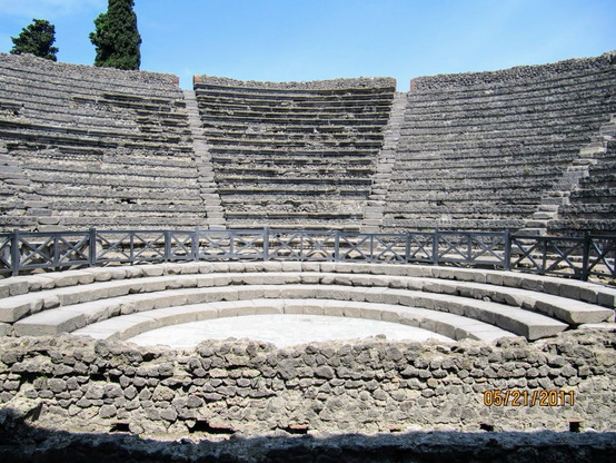 Ruins is a Roman Amphitheater. Circular stone stage on the bottom. Few flight of steps around the stage. A metal barrier around the area. Multiple rows of stone seating rising above the round stage. 