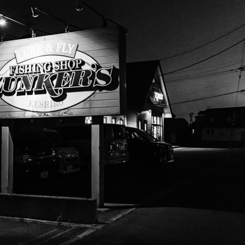 In this black-and-white photograph, a nighttime scene is depicted featuring a storefront and a large sign. The sign is prominently displayed in the foreground on the left side of the image, mounted on two posts. It has a curved top and contains bold lettering. Behind the sign, a building with a pointed roof is visible, housing a shop. The shop is well-lit from the inside, with lights shining through large windows, creating a stark contrast against the dark exterior and surrounding area. To the left of the shop, several parked vehicles can be seen, mostly in shadow, making it difficult to distinguish their specific models or colors.The scene is set against a backdrop of a dark sky, with power lines stretching across the top right portion of the image, adding to the urban feel. The street in front of the shop and the parking area is dimly lit, with some areas in complete darkness, contributing to the overall quiet and somewhat deserted atmosphere of the photograph.