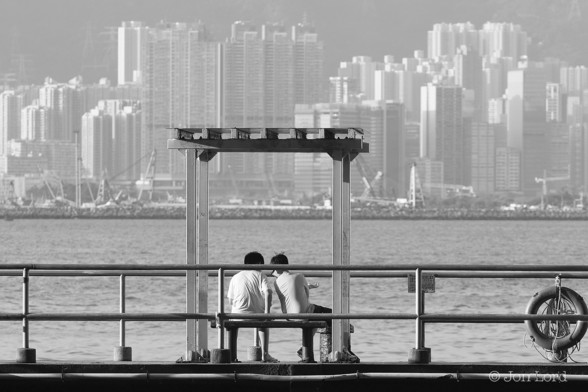 A Black And White Photo In Landscape Format. In The Centre And Foreground Are Two People Sitting On A Small Bench Underneath A Small Rectangular Storm Shelter. The Person On The Right Is Leaning To The Left As If Looking At Something. Stretching Across the Photo Are Two Rows Of Railings. At The Far Right, A Life Buoy Is Attached To The Railing. In The Middle Of The Image Is A Body Of Water. Above Is A Breakwater, Again Stretching From The Left To The Right. Just Above The Breakwater There Are Many Cranes. Beyond: A Great Many White High Rise Apartment Buildings. The Scene Is Set On The Waterfront In North Point, Hong Kong. The Body Of Water Is Victoria Harbour. The High Rise Buildings Are Located In Kowloon City. 