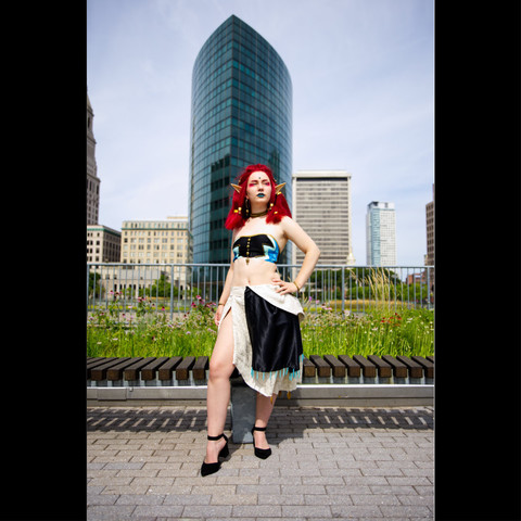 This is a photo of a young woman who is cosplaying as Makeela Riju from the video game Zelda. Her hair is a bright color red and she is dressed in a halter top with a dress that has a slit on the left side. She is in a standing in a confident pose looking upwards and off to the right side. She is standing outdoors and the background displays a downtown city skyline in Hartford Connecticut.