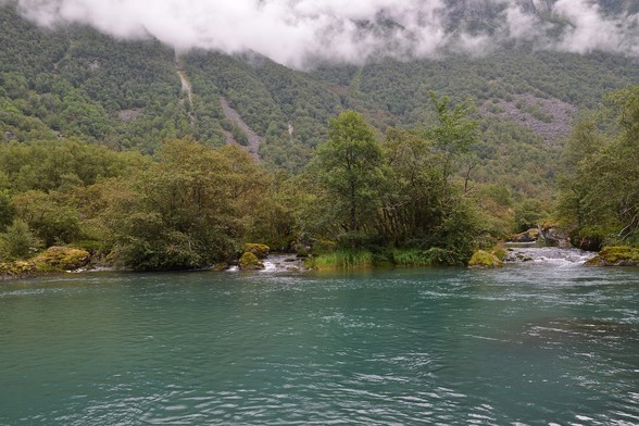 A photo of a river with a forest covered mountainside on the far side. There are low clouds going over the forest. The water in the river is slightly turquoise from a melting glacier.