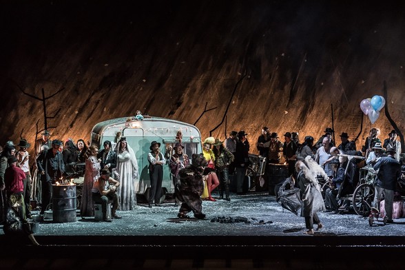 2017年伦敦皇家歌剧院版《游吟诗人》（Il Trovatore）剧照。该版本将舞台背景搬到现代（剧中出现了坦克和机枪）。照片中，中左侧穿婚纱的“大胸女人”是由合唱团的一位男性成员来扮演。