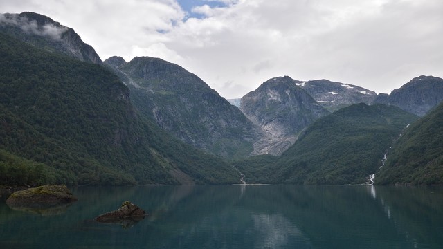 A photo of a lake. There are low mountains on the far side and a glacier melting into the lake from a small waterfall. The lake is slightly turquoise. The sky is cloudy.