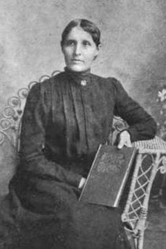 Almeda Jones St. Clair, from a 1909 publication; a Dakota (Sioux) woman wearing a black dress with a high collar, seated in a wicker chair , holding a book