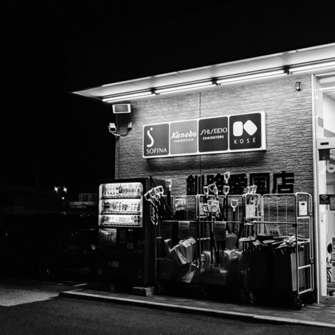 The black-and-white photograph depicts the exterior of a store at night. The building is well-lit from above, casting bright light onto the signage and the area below. The signs prominently display the logos of various cosmetic brands: Sofina, Kanebo, Shiseido, and Kose. Below the signs, there is a vending machine, which is illuminated and filled with various beverages. To the right of the vending machine, there are several shopping carts and a stack of items, including what appear to be snow shovels and other tools, neatly arranged. The store's entrance is partially visible on the far right side of the image, with additional lighting coming from inside the store.