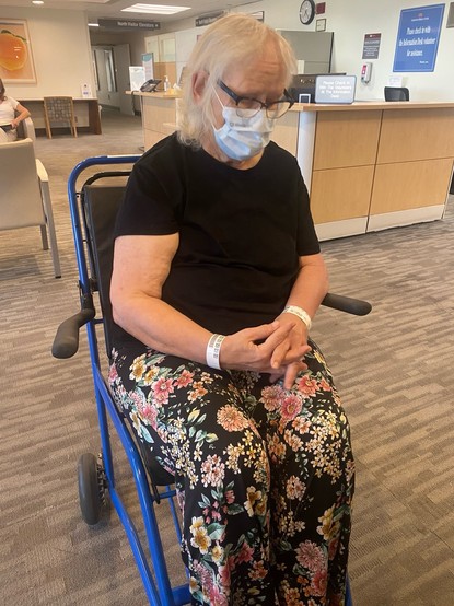 Mom in a black blouse and flower printed slacks in the hospital wheelchair. 
