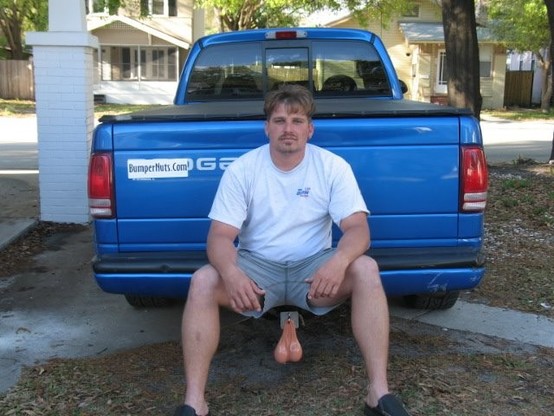 A man sitting on his truck bumper ubove the tow hitch that a plastic pair of testicles are hanging from. 
